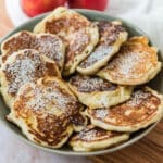 view of grey serving bowl with Polish apple pancakes arranged in a ring around the bowl. Pancakes are dusted with powdered sugar and two apples in background.
