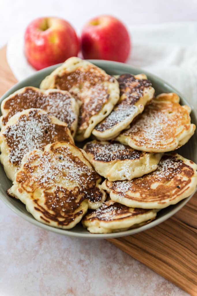 view of grey serving bowl with Polish apple pancakes arranged in a ring around the bowl. Pancakes are dusted with powdered sugar and two apples in background.