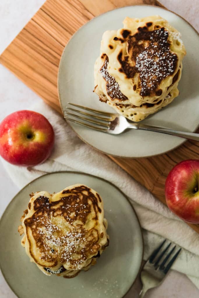 overhead view of two grey plates, each with two thick apple pancakes dusted with powdered sugar, with forks and apples beside